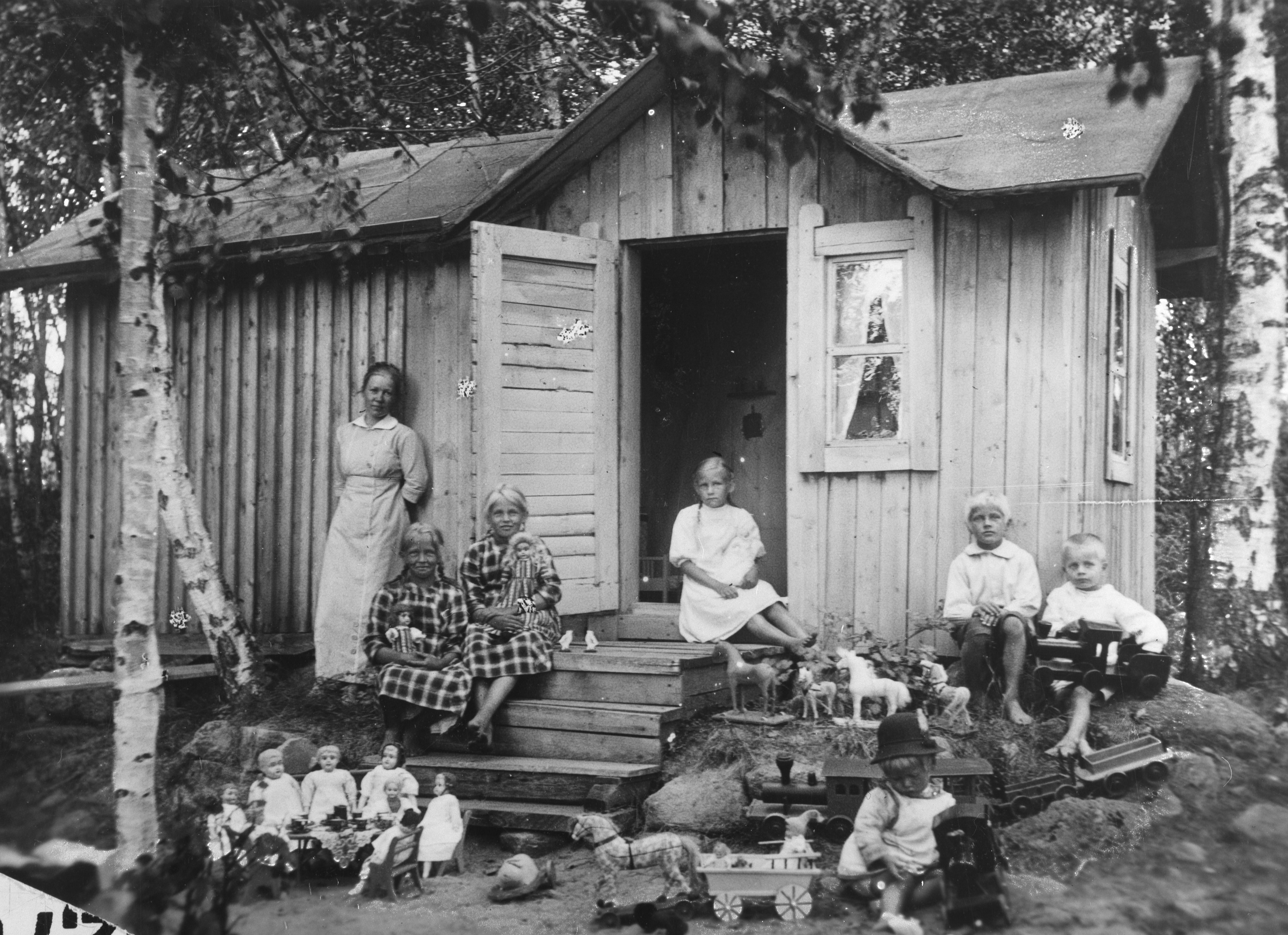 Kuvassa on puutalo. Talon ulkopuolella seisoo nainen vasemmalla puolella. Rappusilla ja talon vasemmalla puolella istuu kuusi lasta. Kuvan etuosassa näkyy leikkikaluja.