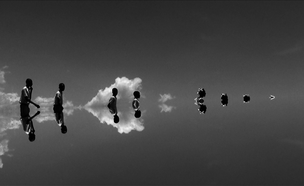 Mustavalkoisessa kuvassa eriasteisesti näkyviä ihmisiä, pilviä ja näiden heijastuksia.