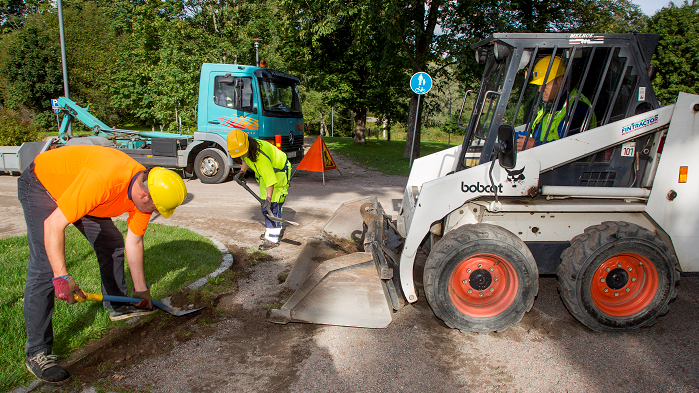 Valokuvassa kaksi työasuista henkilöä lapioi maa-ainesta Bobcat-kuormaajan kauhaan. Taustalla lava-auto, puita ja puistomaisemaa.