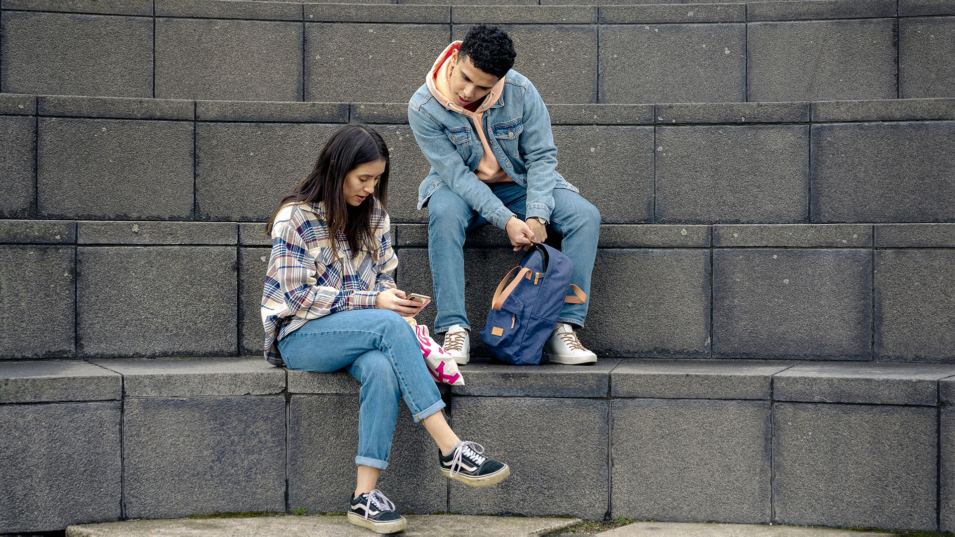 Nuori nainen ja nuori mies istuvat kivirappusilla ja katsovat naisen kädessä olevaa mobiilipuhelinta.
